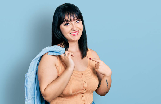 Young hispanic plus size woman wearing holding denim jacket smiling happy pointing with hand and finger  - Foto, Bild