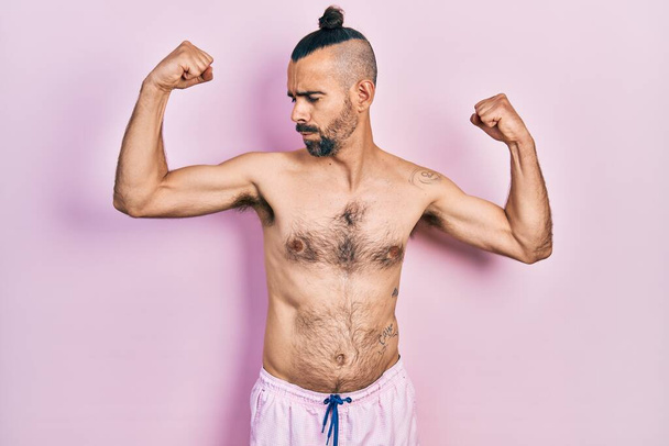 Joven hombre hispano sin camisa usando traje de baño mostrando los músculos de los brazos sonriendo orgulloso. concepto de fitness.  - Foto, imagen