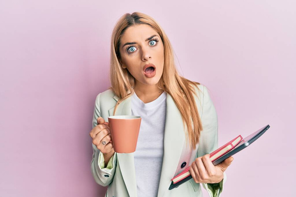 Jonge blanke vrouw drinken van een kopje koffie en houden laptop in shock gezicht, op zoek sceptisch en sarcastisch, verrast met open mond  - Foto, afbeelding