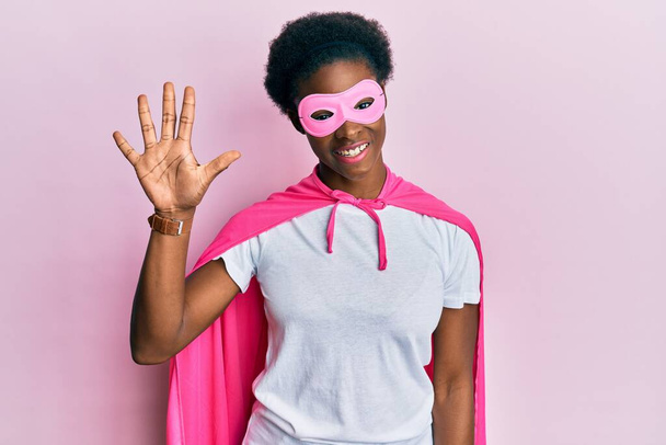 jong Afrikaans amerikaans meisje dragen superheld masker en cape kostuum tonen en wijzen met vingers nummer vijf terwijl glimlachen zelfverzekerd en gelukkig.  - Foto, afbeelding