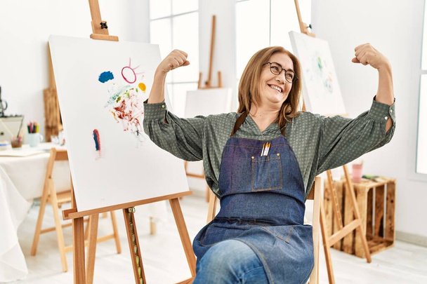 Kobieta w średnim wieku artystka w pracowni sztuki pokazuje mięśnie ramion uśmiechnięte dumą. koncepcja sprawności.  - Zdjęcie, obraz