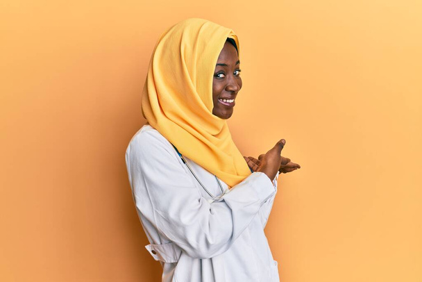 Bella africana giovane donna indossando uniforme medico e hijab invitando a entrare sorridente naturale con la mano aperta  - Foto, immagini