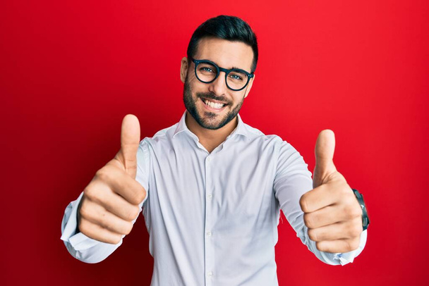 若いヒスパニック系のビジネスマンのシャツや眼鏡を着用し、手で積極的なジェスチャーを行うことを承認し、親指を笑顔と成功のために幸せ。勝者のジェスチャー.  - 写真・画像