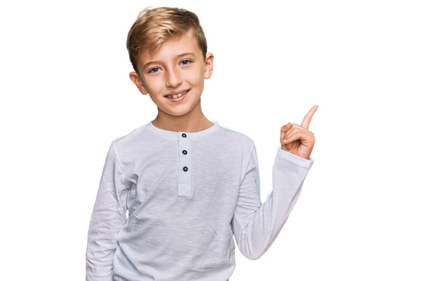 Kleiner kaukasischer Junge in lässiger Kleidung mit einem breiten Lächeln im Gesicht, der mit der Hand und dem Finger zur Seite zeigt und in die Kamera blickt.  - Foto, Bild