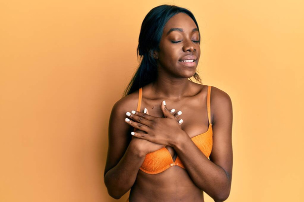 Jovem afro-americana vestindo lingerie sorrindo com as mãos no peito, olhos fechados com gesto grato no rosto. conceito de saúde.  - Foto, Imagem