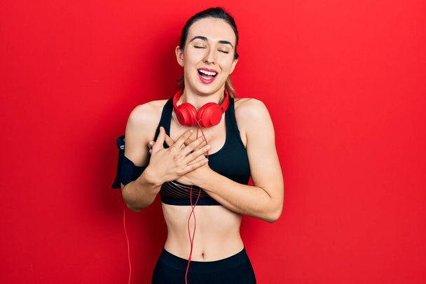 Молода іспаномовна дівчина носить спортивний одяг та використовує навушники, посміхаючись руками на грудях із закритими очима та вдячним жестом на обличчі. концепція здоров'я
.  - Фото, зображення