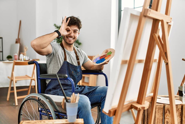若いヒスパニック系の男が車椅子の絵の上に座っているアートスタジオで手の笑顔でokジェスチャーをし、目は幸せな顔で指を通して見る.  - 写真・画像