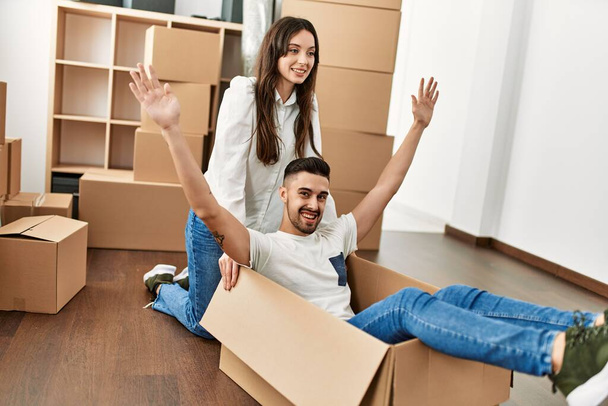 Joven pareja hispana sonriendo feliz jugando usando la caja de cartón como un coche en un nuevo hogar. - Foto, imagen