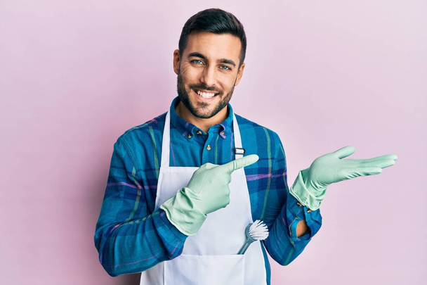 Young hispanic man wearing apron amazed and smiling to the camera while presenting with hand and pointing with finger.  - Foto, Imagem