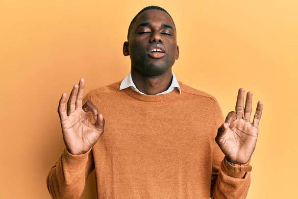 Genç Afro-Amerikan adam günlük kıyafetler giyip rahatlıyor ve gözleri kapalı gülümsüyor parmaklarıyla meditasyon hareketi yapıyor. yoga konsepti.  - Fotoğraf, Görsel