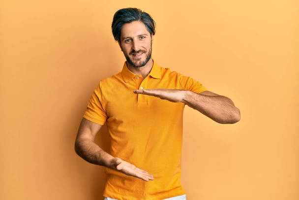 Νεαρός ισπανόφωνος άνδρας φορώντας casual κίτρινο t πουκάμισο gesturing με τα χέρια που δείχνουν μεγάλο και μεγάλο μέγεθος σημάδι, μέτρο σύμβολο. Χαμογελώντας κοιτάζοντας την κάμερα. έννοια μέτρησης.  - Φωτογραφία, εικόνα