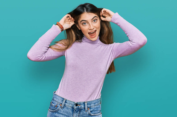 Young beautiful teen girl wearing turtleneck sweater smiling pulling ears with fingers, funny gesture. audition problem  - 写真・画像