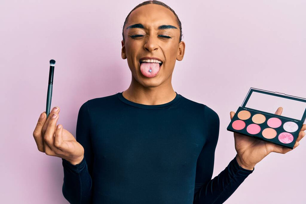 Hispanic man wearing make up and long hair holding makeup brush and blush sticking tongue out happy with funny expression.  - Photo, image