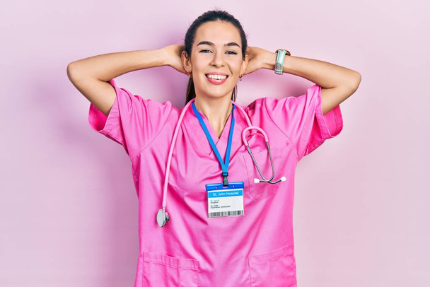 Νεαρή μελαχρινή γυναίκα με στολή γιατρού και στηθοσκόπιο που χαλαρώνει και τεντώνει, χέρια και χέρια πίσω από το κεφάλι και το λαιμό χαμογελώντας ευτυχισμένη  - Φωτογραφία, εικόνα
