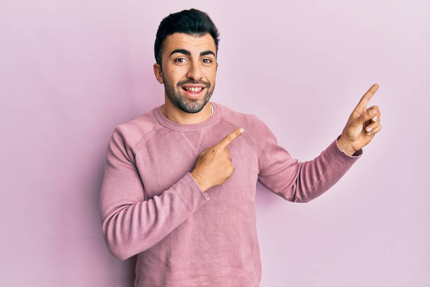 Young hispanic man wearing casual clothes smiling and looking at the camera pointing with two hands and fingers to the side.  - Photo, Image