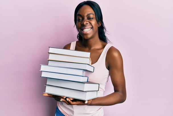 Junge afrikanisch-amerikanische Frau mit Büchern, die zwinkernd in die Kamera blicken, mit sexy Ausdruck, fröhlichem und glücklichem Gesicht.  - Foto, Bild