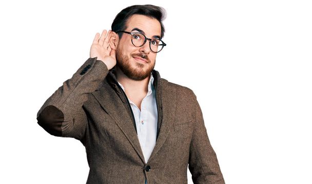 Jonge Spaanse man met een zakenjasje en een bril die glimlacht met hand op oor, luisterend naar geruchten of roddels. doofheid.  - Foto, afbeelding
