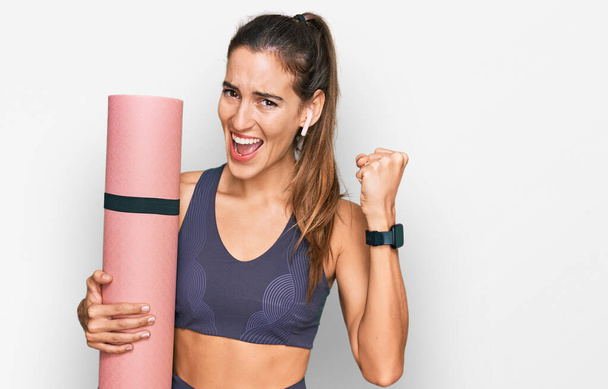 Young beautiful woman wearing sportswear holding yoga mat screaming proud, celebrating victory and success very excited with raised arms  - Photo, Image
