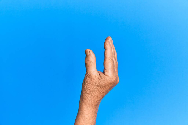 Hand eines hochrangigen hispanischen Mannes über blauem isoliertem Hintergrund, der unsichtbare Dinge pflückt und nimmt, während er mit den Fingern ein Objekt hält, das Raum zeigt  - Foto, Bild