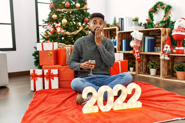 Jeune homme hispanique avec barbe assis près de l'arbre de Noël célébrant 2022 nouvelle année couvrant la bouche avec la main, choqué et effrayé par l'erreur. expression surprise  - Photo, image