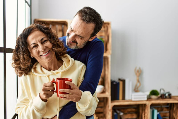 Hispánský pár středního věku se šťastně usmívá a pije kávu. Opírající se o okno doma. - Fotografie, Obrázek
