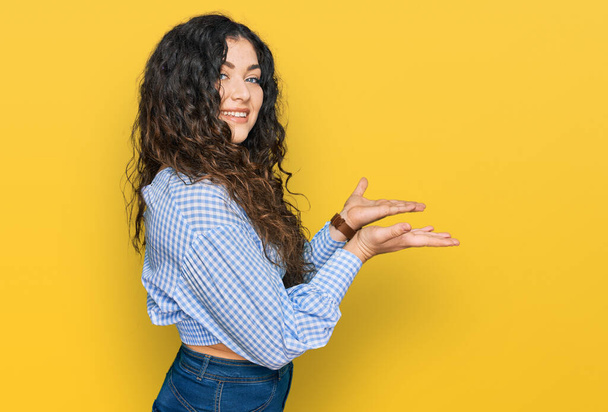 Mladá hispánská dívka na sobě ležérní oblečení ukazuje stranou s rukama otevřenýma dlaněmi ukazující kopírovací prostor, prezentuje reklamu s nadšeným úsměvem  - Fotografie, Obrázek