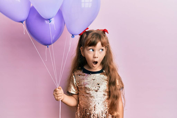 Petite fille caucasienne enfant tenant des ballons peur et étonné avec la bouche ouverte pour la surprise, visage d'incrédulité  - Photo, image