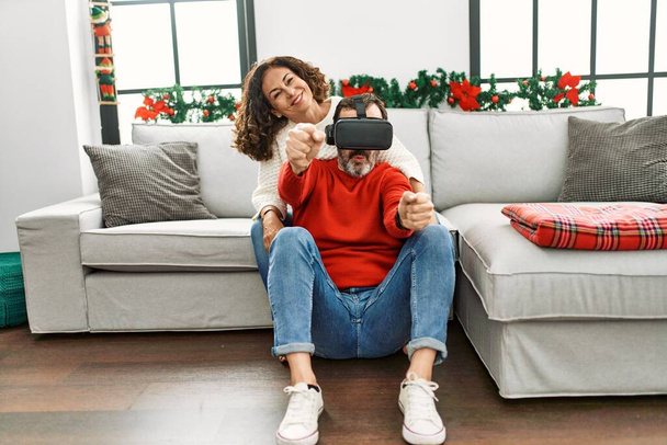 Middle age hispanic couple smiling happy sitting on the sofa playing with virtual reality glasses at home. - 写真・画像