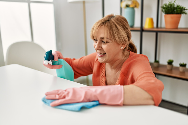 Kobieta w średnim wieku uśmiechnięta szczęśliwa sprzątanie za pomocą dyfuzora i szmaty w domu. - Zdjęcie, obraz