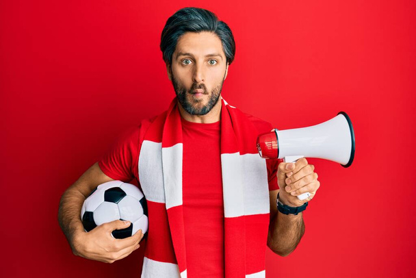 Young hispanic man football hooligan holding ball using megaphone making fish face with mouth and squinting eyes, crazy and comical.  - Photo, Image