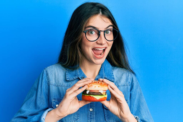 Młoda kaukaska kobieta je smacznego hamburgera świętując szaleństwo i zdumiony sukcesem z otwartymi oczami krzycząc podekscytowany.  - Zdjęcie, obraz