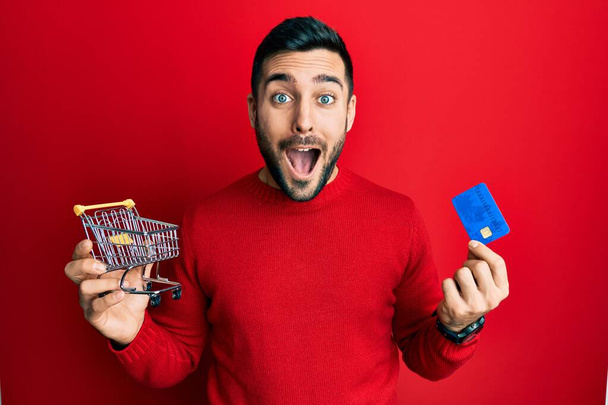 Homem hispânico jovem segurando carrinho de compras pequeno supermercado e cartão de crédito comemorando louco e espantado pelo sucesso com os olhos abertos gritando animado.  - Foto, Imagem