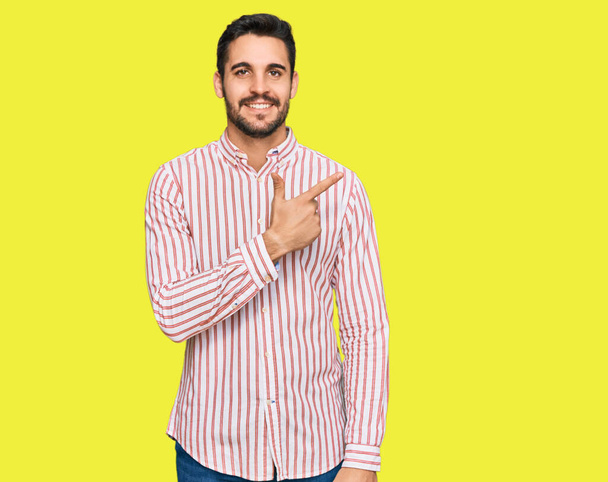Νεαρός Ισπανός που φοράει επαγγελματική μπλούζα χαρούμενος με χαμόγελο στο πρόσωπο δείχνοντας με το χέρι και το δάχτυλο στο πλάι με χαρούμενη και φυσική έκφραση στο πρόσωπο  - Φωτογραφία, εικόνα
