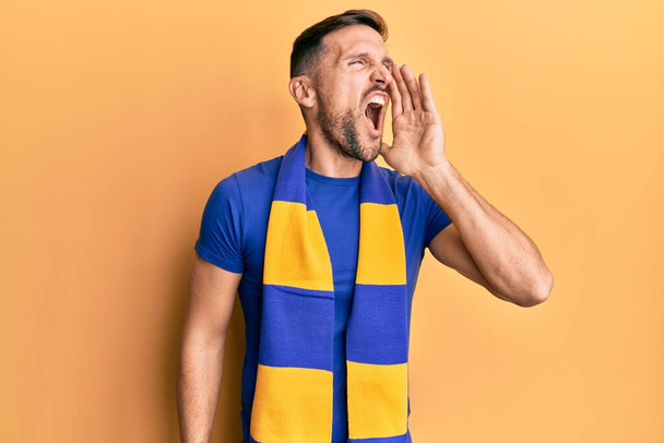Handsome man with beard football hooligan cheering game shouting and screaming loud to side with hand on mouth. communication concept.  - Foto, imagen