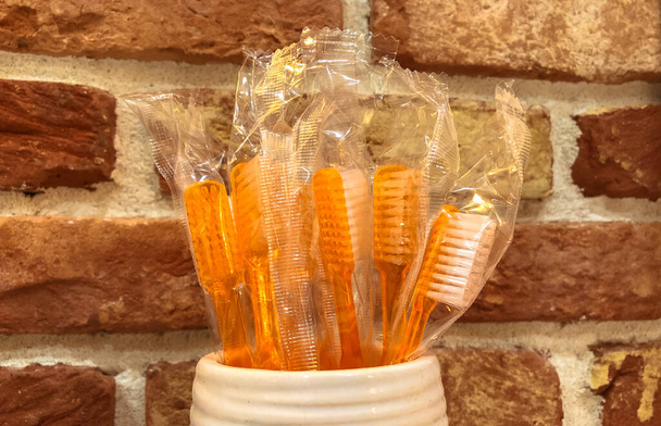 Einweg-Zahnbürsten aus orangefarbenem Kunststoff in einem Keramikbecher gegen eine rote Ziegelwand. Jede Zahnbürste ist einzeln verpackt. Zahnarztbedarf für die Mundpflege. Vorsichtsmaßnahmen gegen Karies. Zahngesundheit - Foto, Bild