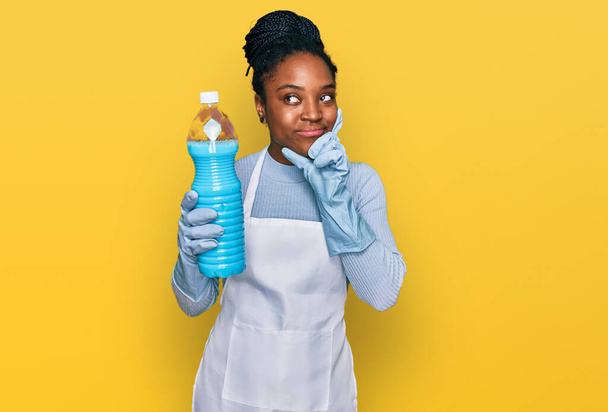 Молода афроамериканська жінка, одягнена в фартух, тримаючи пляшку з миючим засобом серйозно, думає про питання з рукою на підборіддя, роздумуючи про плутанину ідею  - Фото, зображення