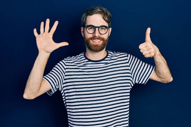 Kaukasischer Mann mit Bart, gestreiftem T-Shirt und Brille, der mit Finger Nummer sechs zeigt und nach oben zeigt, während er selbstbewusst und glücklich lächelt.  - Foto, Bild