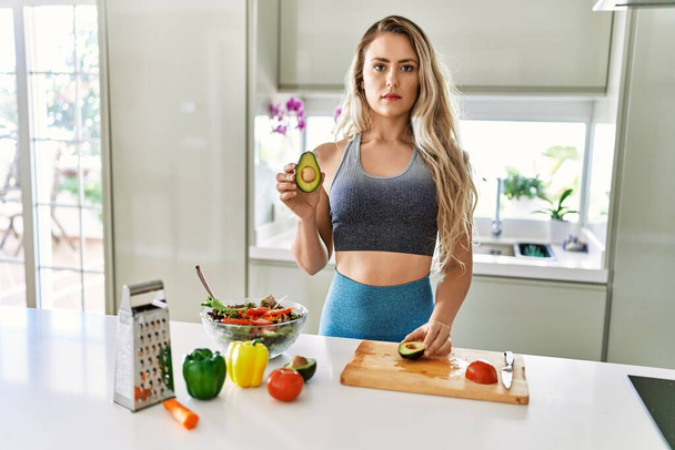 キッチンで健康的なサラダを準備するスポーツウェアを身に着けている若い白人フィットネス女性思考態度と落ち着いた表情自信を持って見える  - 写真・画像