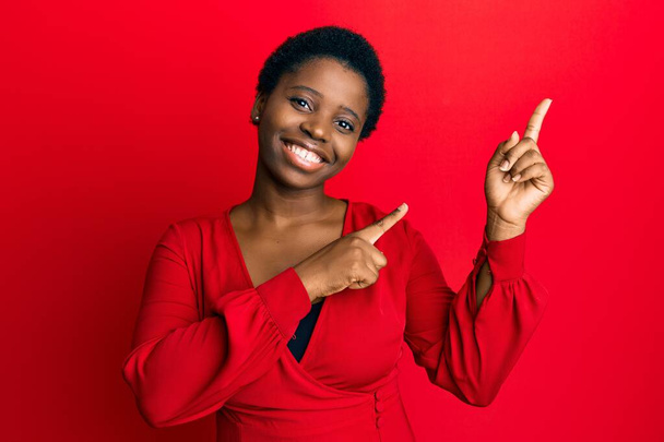 Afro saçlı genç bir kadın kırmızı arka planda günlük kıyafetler giyiyor gülümsüyor ve kameraya bakıyor iki eliyle ve parmaklarıyla kenarı işaret ediyor..  - Fotoğraf, Görsel