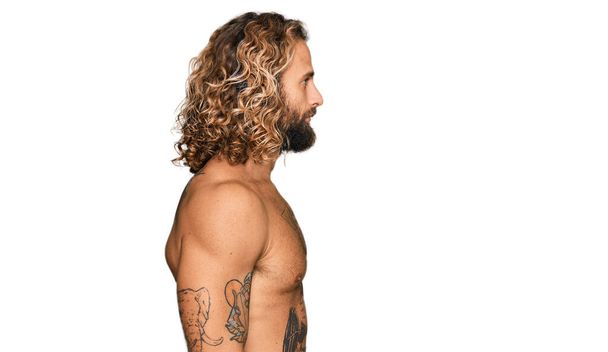 Przystojny mężczyzna z brodą i długimi włosami stojący bez koszulki pokazując tatuaże patrząc na bok, relaks poza profilem z naturalną twarzą i pewny siebie uśmiech.  - Zdjęcie, obraz