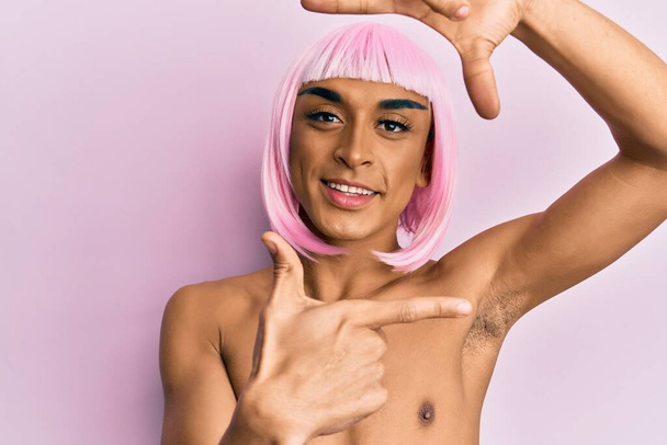 Homem transexual hispânico vestindo maquiagem e peruca rosa sorrindo fazendo moldura com mãos e dedos com rosto feliz. conceito de criatividade e fotografia.  - Foto, Imagem