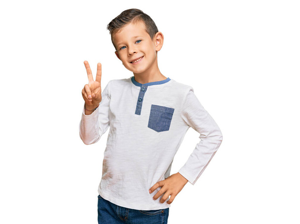 Imádnivaló kaukázusi gyerek alkalmi ruhát visel, mutatva és mutogatva a kettes számú ujjaival, miközben magabiztosan és boldogan mosolyog..  - Fotó, kép