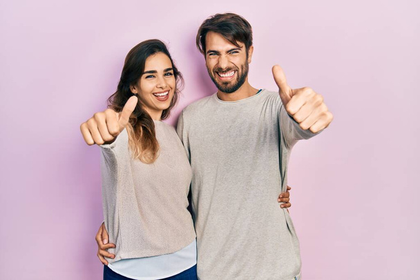 Молодая испаноязычная пара в повседневной одежде, одобряющая позитивный жест рукой, улыбается и радуется успеху. жест победителя.  - Фото, изображение