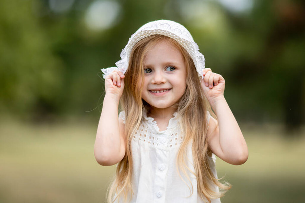 избирательный фокус. Милая маленькая девочка, блондинка с длинными волосами, улыбается, надевает шляпу в парке. Жара, ходьба, предотвращение солнечных ударов, обезвоживание. Летние белые люки, шляпа из панамы, защита от солнца - Фото, изображение