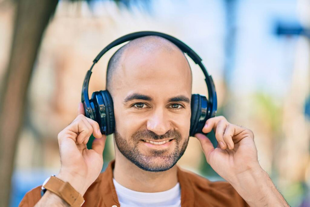 Fiatal spanyol kopasz férfi mosolyog boldogan használ fejhallgatót a városban - Fotó, kép