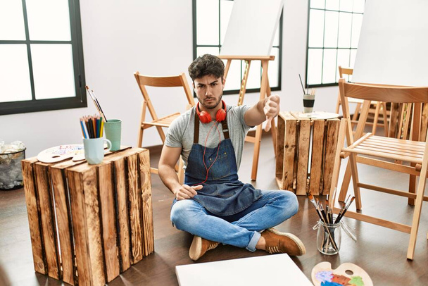 Młody Hiszpan siedzący w pracowni artystycznej, nieszczęśliwy i wściekły, okazujący odrzucenie i negatyw z opuszczonymi kciukami. złe wyrażenie.  - Zdjęcie, obraz