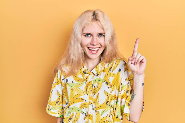 Belle femme caucasienne aux cheveux blonds portant une chemise colorée pointant du doigt avec une idée réussie. Sorti et heureux. numéro un.  - Photo, image