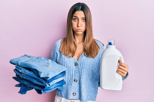 Brunette young woman holding jeans for laundry and detergent bottle puffing cheeks with funny face. mouth inflated with air, catching air.  - Фото, зображення