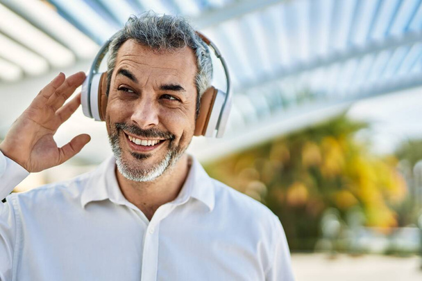 Středního věku šedovlasý muž s úsměvem šťastný pomocí sluchátek ve městě. - Fotografie, Obrázek