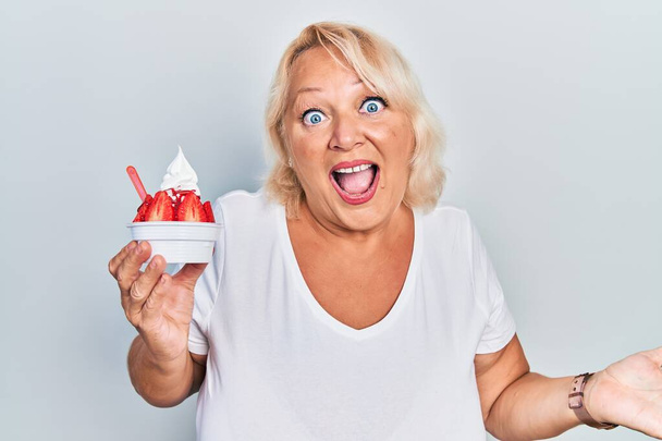 Középkorú szőke nő eszik eper fagylalt ünneplő eredmény boldog mosollyal és a győztes kifejezés emelt kézzel  - Fotó, kép